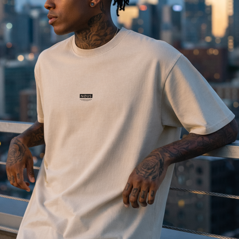 Man with tattoos wearing a beige t-shirt against a city skyline at dusk