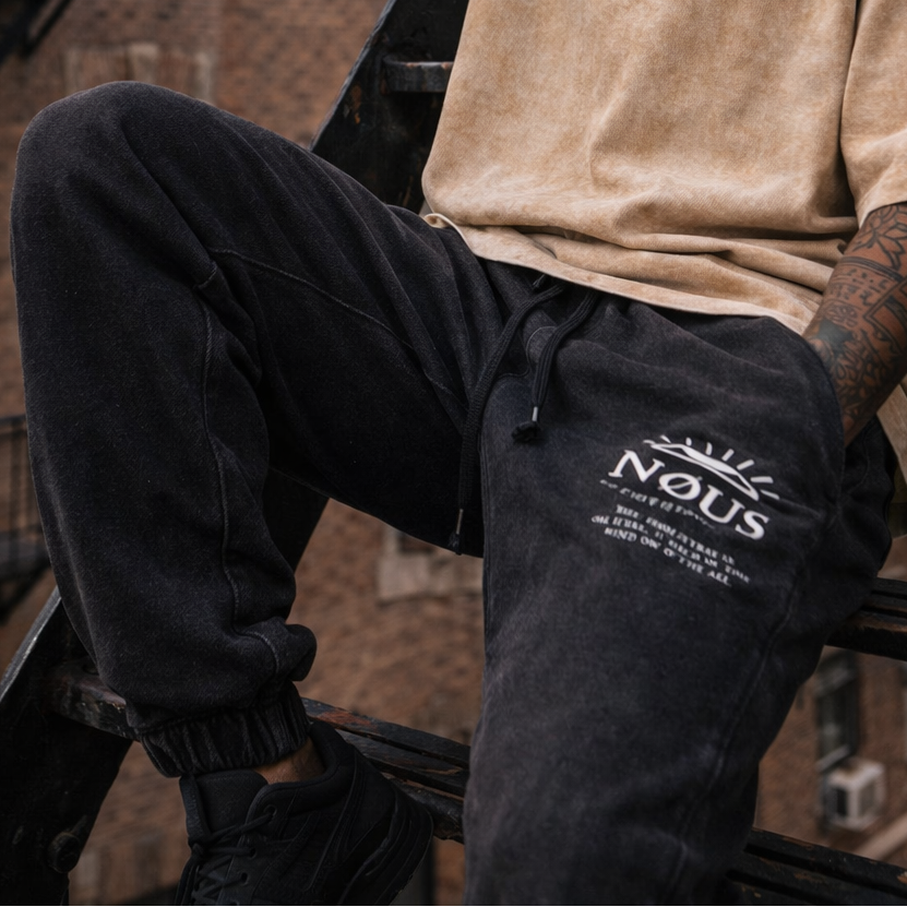 Man sitting on a fire escape wearing a beige t-shirt and black pants with text.