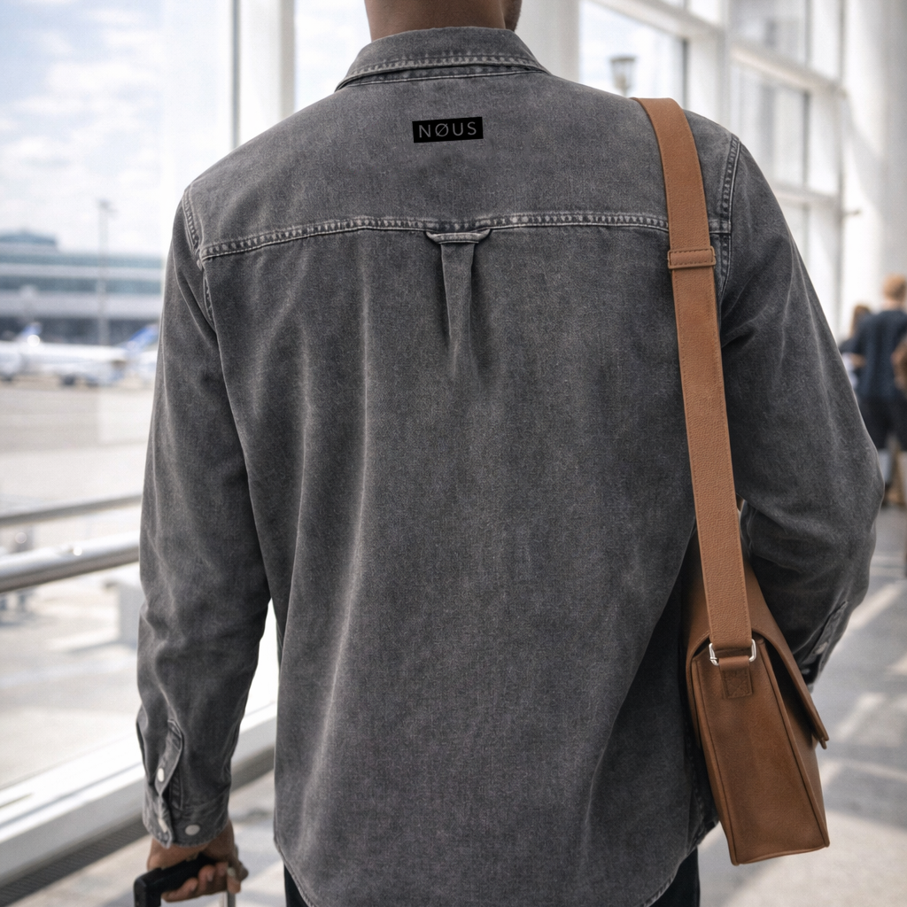 Man in a gray denim shirt with a brown leather bag walking through an airport terminal.