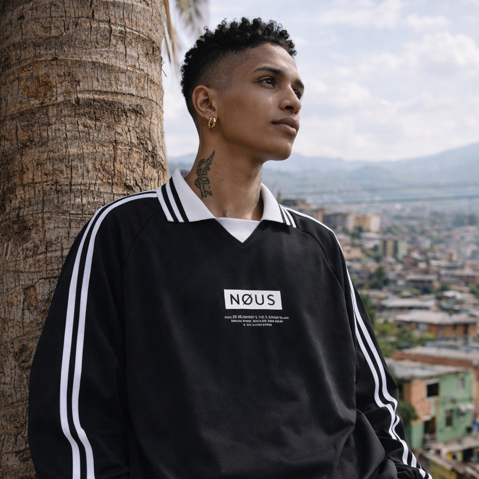 Person wearing a black tracksuit with 'NOUS' logo, leaning against a palm tree with a cityscape in the background.