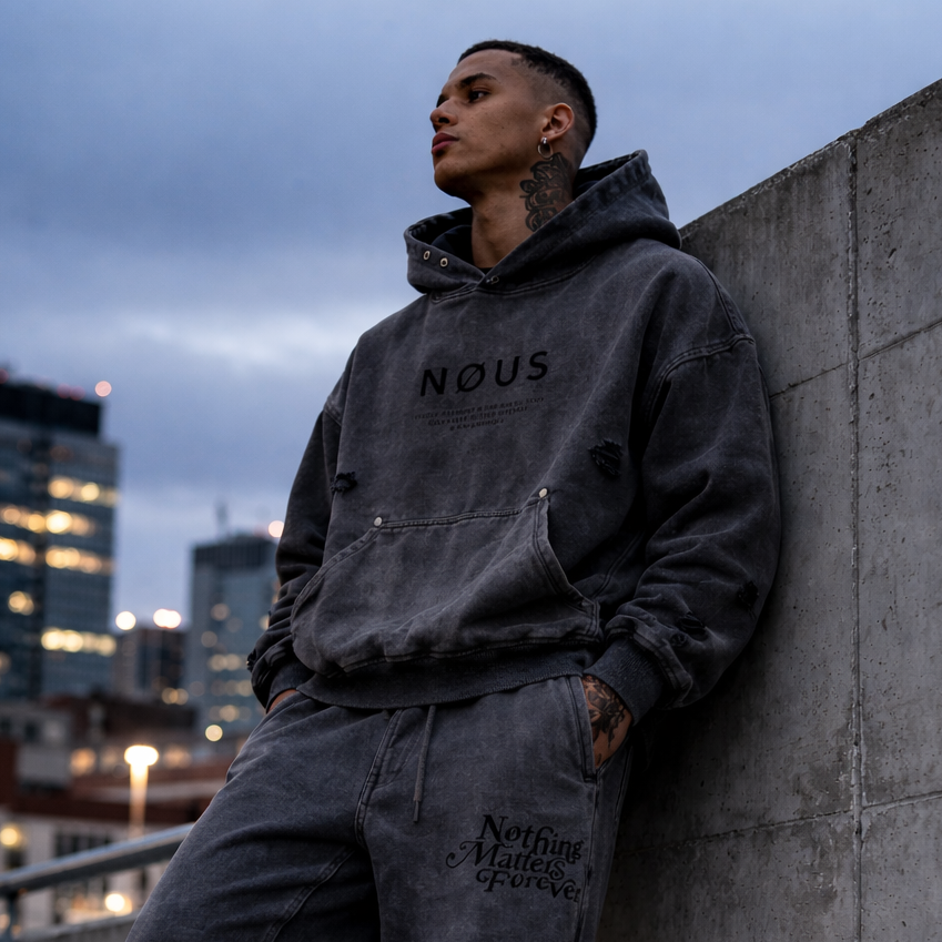 Person wearing a dark hoodie and pants leaning against a concrete wall with cityscape in the background