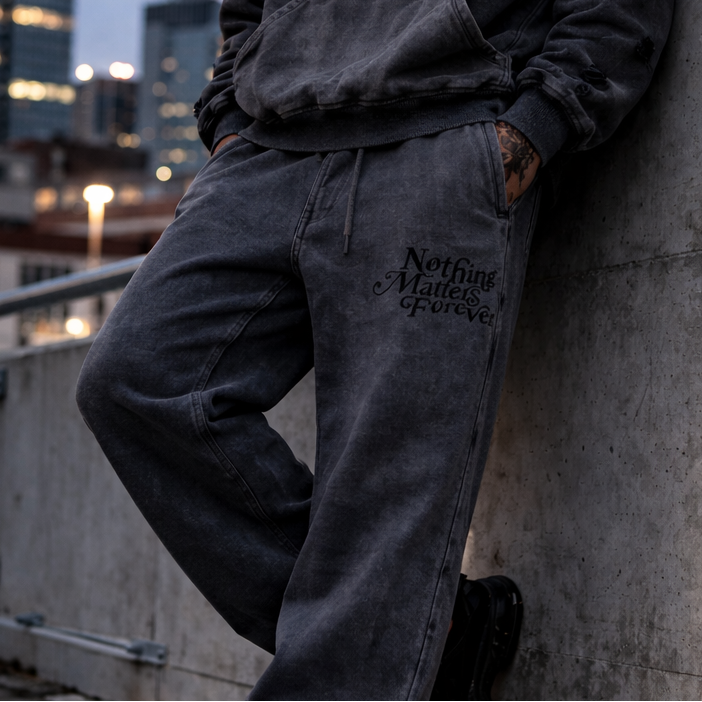 Person wearing a dark hoodie and pants leaning against a concrete wall with cityscape in the background