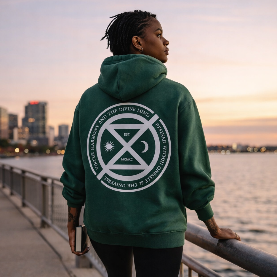 Person wearing a green hoodie with a visible logo, standing by a waterfront at sunset.