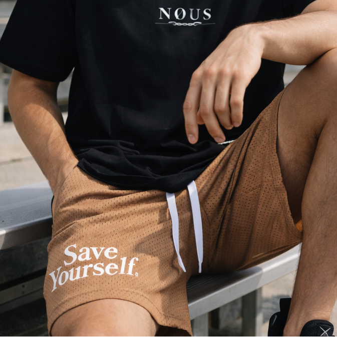 Man sitting on bleachers wearing a black t-shirt with 'NOUS' and brown shorts with 'Save Yourself' text.